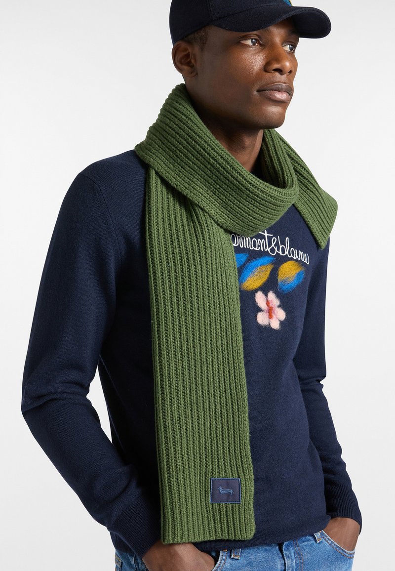 Groene geribbelde sjaal van gebreid materiaal, om de nek gedrapeerd, met een klein logopatch. Draagt een donkere trui en een pet.