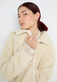 Beige, fluffig sherpa-jacka med krage, hög halsringning och ribbade ärmslut. Modellen håller i kragen och visar upp den mjuka texturen.