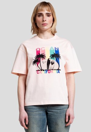 Kvinde iført lyserød t-shirt med farverigt "Ocean California"-tekst, palmer, surfbrætter og fugle grafik, kombineret med blå jeans.