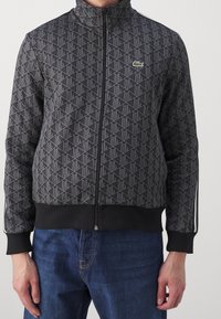 Homme portant une veste noire à motifs avec fermeture éclair, un petit logo crocodile vert et un jean bleu, debout devant un fond clair uni.