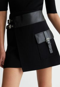 Černá sukně má hladký kožený pas s dvěma stříbrnými přezkami, boční kapsu s detailem přezky a měkkou texturovanou látku.