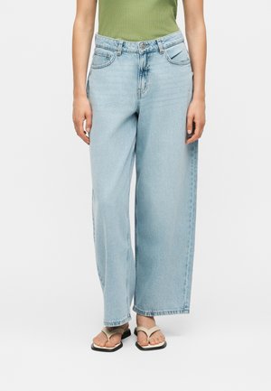 Osoba nosząca jasnoniebieskie szerokie jeansy, zielony bezrękawnik i beżowe klapki, stojąca na białym tle.