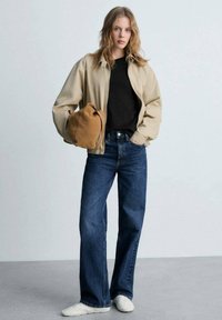 Giacca beige con zip, t-shirt nera, jeans a gamba larga blu scuro e sneakers bianche. Tiene una borsa marrone. Design semplice e vestibilità casual.