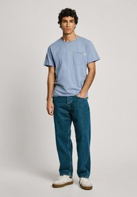 Šviesiai mėlynas medvilninis marškinėlis su krūtinės kišenėle, derinamas su tamsiai mėlynomis džinsinėmis kelnėmis ir baltomis sportinėmis bateliais su rudo atspalvio akcentais.