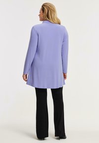 Kvinde med blondt hår iført en langærmet lavendel tunika og sorte bukser, stående og vendt væk mod en ensfarvet baggrund.