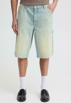 Mann trägt hellblaue Jeansshorts, weiße Kniestrümpfe, schwarze Loafers und ein beigefarbenes Oberteil, die Hände in den Taschen, steht vor einem weißen Hintergrund.