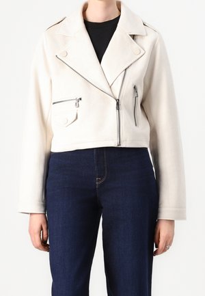 Kobieta ubrana w krótki, kremowy motocyklowy kurtkę z srebrnymi zamkami i ciemnoniebieskie jeansy z wysokim stanem na jednolitym tle.