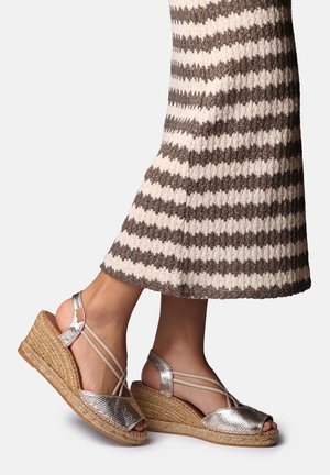 Benedenlichaam gekleed in een bruin-wit gestreepte, gebreide rok en zilverkleurige metalen sleehak espadrille sandalen met gevlochten touwen zolen.