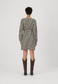 Eine Frau steht mit dem Rücken zur Kamera und trägt ein schwarzes und beige geometrisch gemustertes Kleid mit langen Ärmeln sowie braune Stiefel bis zur Mitte der Wade.