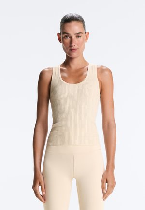 Kobieta z włosami związanymi do tyłu, ubrana w bezrękawnik z beżowego prążkowanego materiału i pasujące legginsy z wysokim stanem, stojąca przodem na białym tle.
