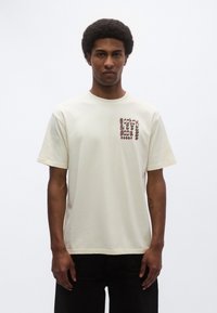 Μπλουζάκι από βαμβάκι σε κρεμ απόχρωση με στρογγυλή λαιμόκοψη και κοντά μανίκια· διαθέτει ένα σχεδιαστικό μοτίβο στο αριστερό στήθος.