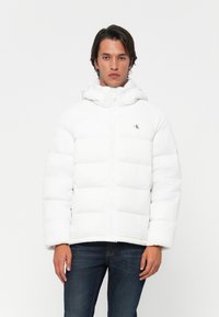 Man in een witte gewatteerde jas met capuchon en Calvin Klein-logo, gecombineerd met donkerblauwe jeans, staand tegen een witte achtergrond.