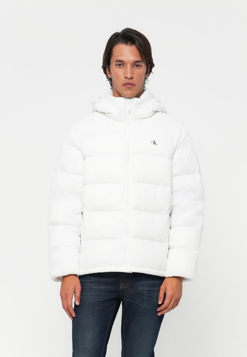 Man in een witte gewatteerde jas met capuchon en Calvin Klein-logo, gecombineerd met donkerblauwe jeans, staand tegen een witte achtergrond.