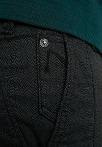 Close-up van een zwarte getextureerde broekzak met een metalen knoop en een groene stoffen tailleband erboven.