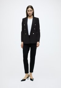 Μαύρο σακάκι διπλής σειράς με ασημένια κουμπιά, φορεμένο πάνω από λευκή μπλούζα, συνδυασμένο με στενά μαύρα παντελόνια και ψηλοτάκουνα πέδιλα με μύτη.