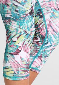 Zbliżenie kolorowych szortów w tropikalny wzór z tealowymi detalami na szwie, na udzie osoby.