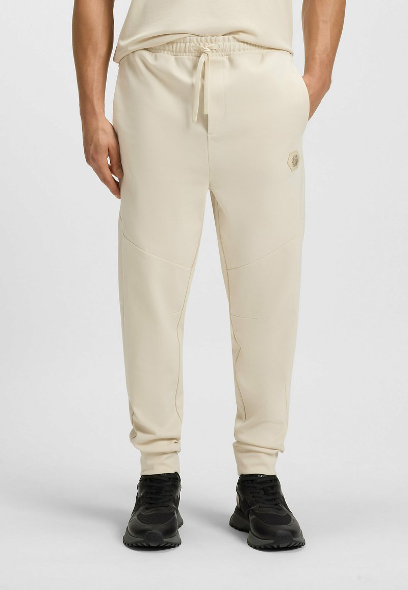 Beige Jogginghosen aus weichem Stoff, mit einem Kordelzugbund, schmal zulaufenden Beinen und seitlichen Taschen. Ein kleines Logo ist an der Seite sichtbar.
