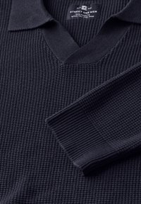 Mörkblå stickad tröja med V-ringning, texturerad tyg, ribbad krage och manschetter, samt en vävd etikett på insidan av halsen.