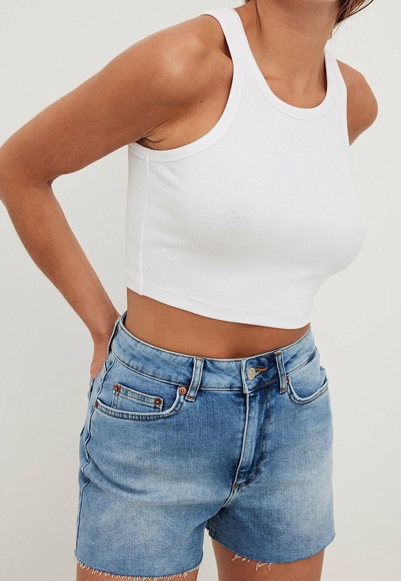 Top corto de tirantes blanco acanalado, combinado con pantalones cortos de denim azul claro, que cuentan con un dobladillo deshilachado y dos bolsillos frontales con remaches de cobre.
