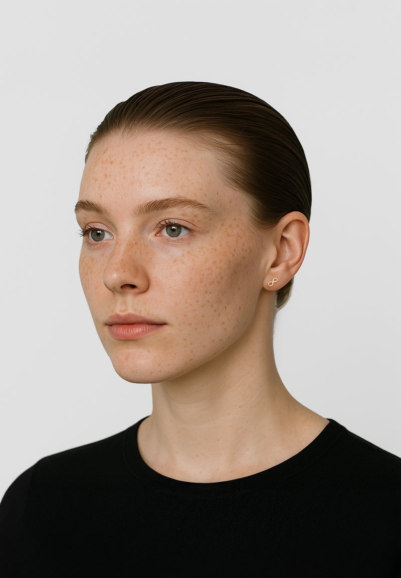 Goldfarbene Ohrringe in Form einer Acht, getragen von einer Person mit heller Haut und Sommersprossen, die ein schwarzes Hemd trägt. Glatte Textur, minimalistisches Design.