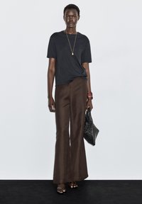 Sort t-shirt med korte ærmer, brune vidde bukser, holder en sort vævet taske med struktur, accessoriseret med en guldfarvet vedhængskæde og en bordeauxrød armbånd.