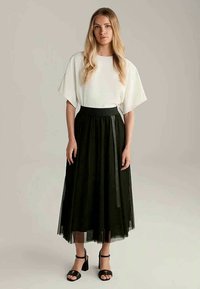 Mulher loira vestindo uma blusa branca de mangas largas, saia midi de tule preta e sandálias de salto pretas, de pé contra um fundo liso.