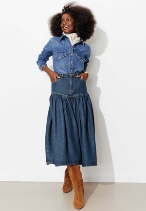 Femme souriante avec afro portant une chemise en jean boutonnée, une longue jupe en jean, une écharpe crème et des bottes montantes en daim marron, debout contre un mur blanc.