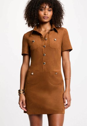 Donna con capelli ricci che indossa un vestito marrone a maniche corte con bottoni e tasche frontali, in piedi davanti a uno sfondo bianco semplice.