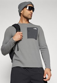 Siva košulja dugih rukava s teksturiranim materijalom, s tamnosivim džepom na prsima i logotipom The North Face. Uključeni su crni ruksak i kapa.