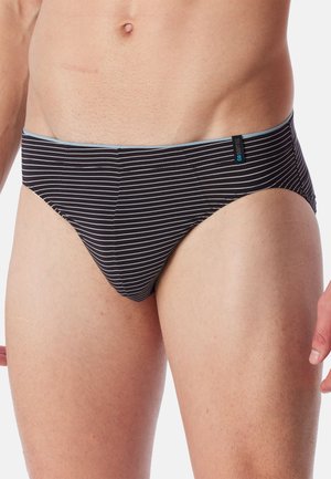 Männlicher Oberkörper, der schwarze Slips mit dünnen weißen horizontalen Streifen und einem kleinen schwarzen Etikett am Bund vor weißem Hintergrund trägt.
