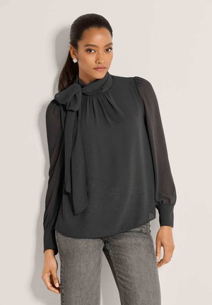 Vrouw met donker haar in een paardenstaart, die een zwarte blouse met lange mouwen en een strik bij de hals draagt en grijze spijkerbroek, kijkt recht vooruit.