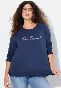 Felpa blu navy con polsini e orlo a scallop, caratterizzata dal testo "New Casual" in corsivo bianco. Il materiale sembra morbido e confortevole.