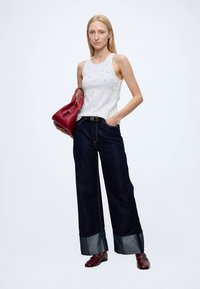 Femme aux longs cheveux blonds portant un haut blanc sans manches, un jean large foncé avec des revers, des chaussures rouges et tenant un sac à main rouge.