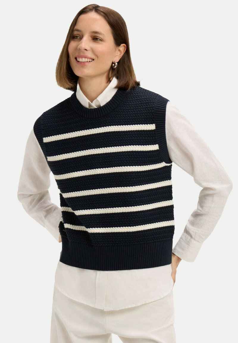 Femme souriante aux cheveux bruns mi-longs portant un gilet en maille bleu marine à rayures horizontales blanches par-dessus une blouse blanche.