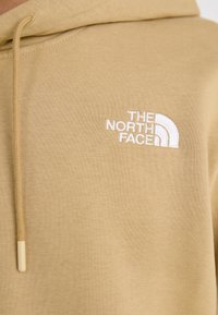 Geltonas gaubtas pagamintas iš minkštos medžiagos, su balta Siuvinėta "The North Face" logotipu kairiajame krūtinės pusėje ir su raišteliu ant gaubto.