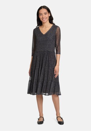 Kleid mit V-Ausschnitt und halbtransparenten Dreiviertelärmeln, marineblaue Stoff mit leichtem floralem Muster; geraffter, gestufter Rock. Schwarze flache Schuhe.