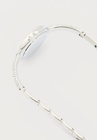 Ασημένιο ρολόι χειρός με στενό, καμπυλωτό λουράκι διακοσμημένο με μικρές διάφανες κρυστάλλινες λεπτομέρειες και στις δύο πλευρές κοντά στο καντράν.