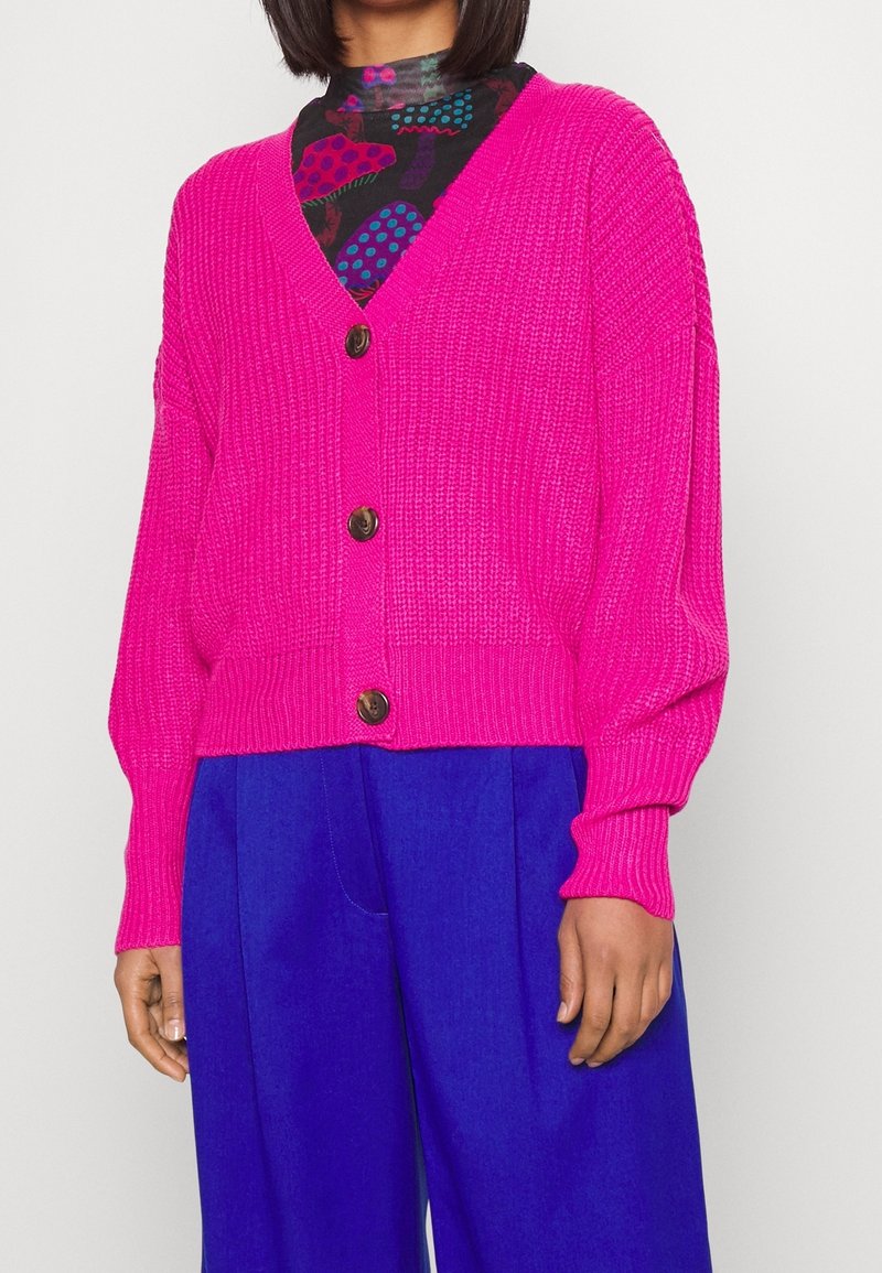 Cardigan rose tricoté avec encolure en V, boutons marron et texture côtelée. Associé à un pantalon bleu à jambes larges.