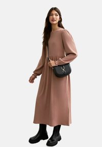 Robe mauve longue à côtes avec manches bouffantes, taille cintrée et sac bandoulière noir. La mannequin porte des bottes noires à semelle épaisse. Fond uni.