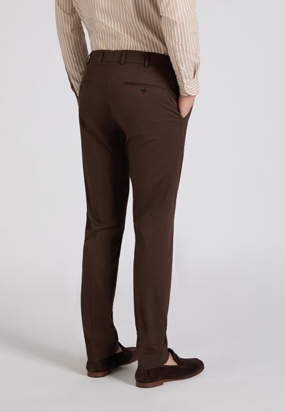 Homme portant un pantalon sur mesure marron foncé, une chemise beige à rayures blanches et des mocassins marron foncé, debout les mains dans les poches.