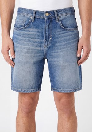 Hombre con pantalones cortos de mezclilla azul de longitud media con bolsillos delanteros y bragueta con botones, de pie con las manos relajadas a los lados.