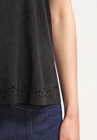 Haut noir sans manches avec un détail floral découpé au laser sur l'ourlet, porté avec un jean en denim bleu foncé, montrant le bras droit et le côté du torse.