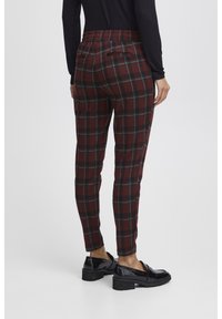 Pantaloni a quadri bordeaux con accenti verdi e neri, dotati di una cintura elastica e tasche posteriori, abbinati a mocassini in vernice nera.