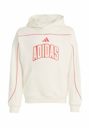 Persona joven con sudadera blanca de Adidas, pantalones deportivos rojos de Adidas, gorra roja y zapatillas blancas, de pie con las manos en los bolsillos.