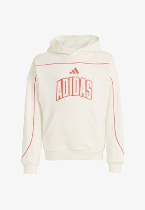Persona joven con sudadera blanca de Adidas, pantalones deportivos rojos de Adidas, gorra roja y zapatillas blancas, de pie con las manos en los bolsillos.