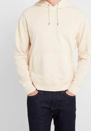 Mann trägt einen hellbeigen Hoodie mit Kängurutasche und Kordeln, kombiniert mit dunkelblauen Jeans, Hände in den Taschen.