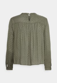 Blusa verde oliva a maniche lunghe in tessuto testurizzato, con dettaglio di apertura a chiave sul retro, pieghe verticali e piccoli punti in rilievo su tutta la superficie.