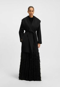 Black wrap coat with wide collar, belted waist, and long sleeves, paired with a textured black tiered skirt, creating a layered look.