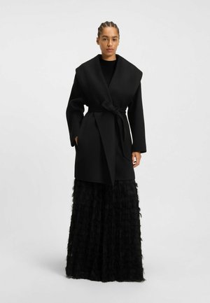 Black wrap coat with wide collar, belted waist, and long sleeves, paired with a textured black tiered skirt, creating a layered look.