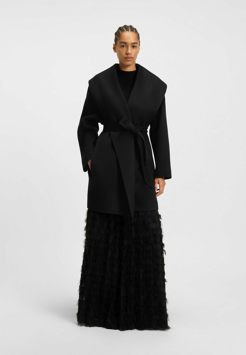 Black wrap coat with wide collar, belted waist, and long sleeves, paired with a textured black tiered skirt, creating a layered look.